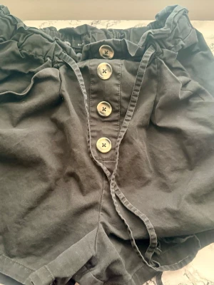 Svarta högmidjade shorts med knappar - Säljer ett par svarta högmidjade shorts med fyra stora dekorativa knappar framtill och ett knytband i midjan. Shortsen har en papperspåseliknande midja och är tillverkade i ett mjukt bomullstyg. Perfekta för varma dagar!