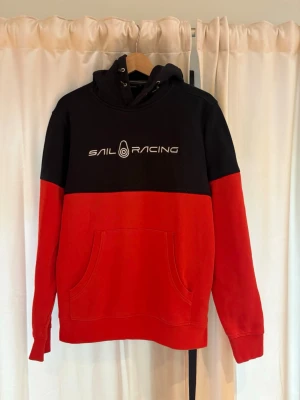 Svart och röd hoodie Sail Racing - Snygg hoodie från Sail Racing i svart och rött med logga framtill. Hoodien har en stor känguruficka, dragsko i huvan och är tillverkad i mjukt bomullsmaterial. Perfekt för en avslappnad och sportig stil.