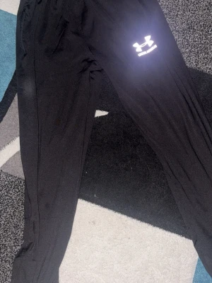 Svarta mjukisbyxor från Under Armour - Svarta mjukisbyxor från Under Armour med resår i midjan och en vit logga på låret. Byxorna har en avslappnad passform och är tillverkade i ett mjukt och stretchigt material som passar perfekt för chill eller träning.