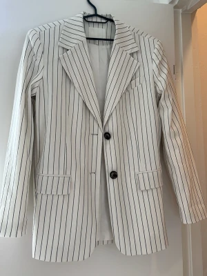 Vit kavaj med svarta ränder - Snygg vit kavaj med smala svarta vertikala ränder och två knappar framtill. Klassisk krage och två fickor med lock. Perfekt för en trendig och stilren look. Rak passform och helfodrad insida.