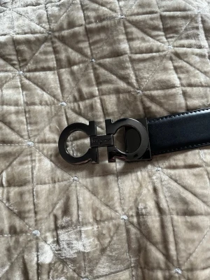 Svart skärp från Salvatore Ferragamo - Snyggt svart skärp från Salvatore Ferragamo med ikoniskt metallspänne i mörk silverfärg. Bältet är tillverkat i slätt skinn. Perfekt accessoar för dig som gillar stilrena detaljer. Använd väldigt fåtal gånger så skärpet är som nytt 