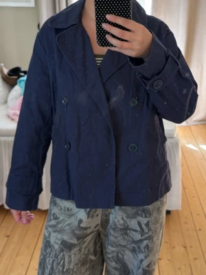 Mörkblå kappa - Stilren mörkblå kappa med bred krage. Jackan har långa ärmar och stora knappar framtill. Passar perfekt för dig som vill ha en klassisk look med modern touch. Materialet ser ut att vara bomull eller en bomullsblandning.