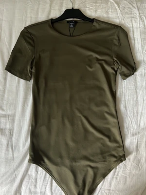 Olivgrön kortärmad body - En stilren olivgrön body med korta ärmar och rund halsringning. Tillverkad i ett mjukt och stretchigt material som sitter skönt mot kroppen. Perfekt att matcha med jeans eller kjol för en enkel och snygg look.
