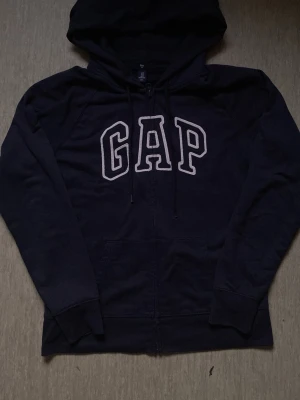 Mörkblå GAP hoodie med dragkedja - Snygg mörkblå hoodie från GAP med vit broderad logga på bröstet. Tröjan har huva med dragsko, långa ärmar och praktiska fickor framtill. Perfekt för en avslappnad och cool stil.