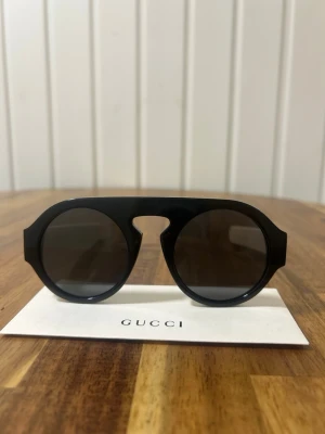 Svarta runda solglasögon från Gucci - Snygga svarta solglasögon från Gucci med rund form och bred båge. Glasögonen har mörka linser och en unik design med markerad brygga. Kommer med originalfodral i sammet. Perfekta för dig som vill sticka ut med en trendig accessoar. 