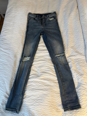 Blå slitna skinny jeans med hål - Säljer ett par blå skinny jeans med slitningar och hål på båda knäna. Jeansen har klassisk femficksmodell, normal midja och är gjorda i stretchigt denimtyg för en tajt passform. Perfekta för dig som gillar en trendig och avslappnad stil.