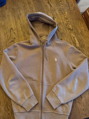 Beige hoodie från Polo Ralph Lauren - Snygg beige hoodie från Polo Ralph Lauren med dragkedja och huva. Klassisk broderad logga på bröstet och ribbade muddar vid ärmslut och nederkant. Perfekt för en avslappnad och stilren look. Ny pris 1500kr 
