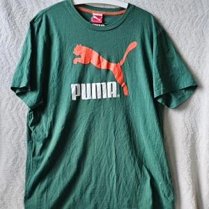 Grön Puma t-shirt med logga - Säljer en grön t-shirt från Puma med stor orange logga och text på bröstet. Klassisk rund halsringning och korta ärmar. Tillverkad i mjuk bomull som känns skön mot huden. Perfekt för dig som gillar sportig och avslappnad stil.