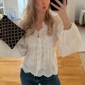 Vit broderad blus med puffärm - Söt vit blus med broderade blommor och romantiska detaljer. Blusen har v-ringning, knytband framtill och vida puffiga långärmar. Materialet är lätt och luftigt, perfekt för en trendig och feminin look.