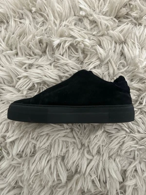 Svarta sneakers i mocka - Stilrena svarta sneakers i mocka med låg profil och tjock svart sula. Skorna har minimalistisk design utan synliga snören och en mjuk ovandel i mocka som ger en lyxig känsla. Perfekta för dig som gillar en clean och modern look.
