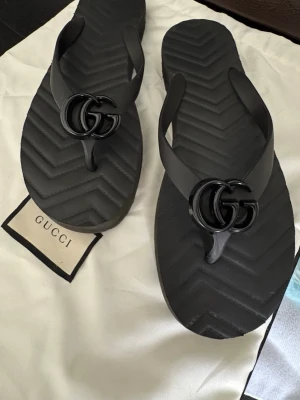 Oanvända flip-flops från Gucci - Ett par svarta och ett par beiga exklusiva flip-flops från Gucci i gummi med den ikoniska GG-loggan framtill. Sandalerna har en stilren design med räfflad sula och breda remmar. Perfekta för sommaren och ger en lyxig touch till din outfit. Priset är för båda två. Orginalpris är 6000kr/st