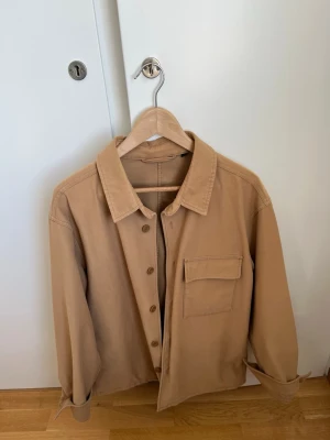 Beige overshirt med ficka - Stilren vårig/höstig beige overshirt med klassisk krage och knappstängning framtill. En bröstficka med lock ger en snygg detalj. Perfekt att bära öppen eller stängd för en avslappnad look.