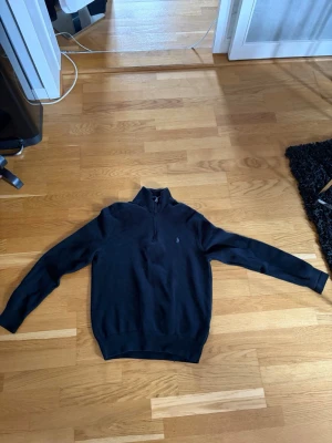 Svart stickad tröja med halv dragkedja - Snygg svart stickad tröja från Polo Ralph Lauren med halv dragkedja och broderad logga på bröstet. Tröjan har hög krage och långa ärmar, perfekt för lager på lager. Materialet känns mjukt och bekvämt.