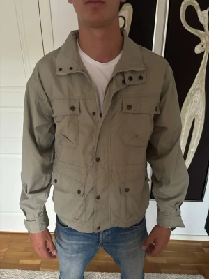 Beige fieldjacket - Snygg beige vindjacka med flera framfickor och tryckknappar. Jackan har hög krage, dragkedja och knappslå framtill. Perfekt för dig som vill ha en stilren och funktionell jacka till vardags. Passar till jeans och t-shirt för en avslappnad look.