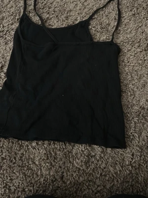 Svart ribbad linnetopp - Svart ribbad linnetopp med smala axelband och enkel design. Toppen är ärmlös och har en tight passform som passar perfekt till jeans eller kjol. Perfekt basplagg för din garderob.