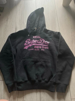 Svart Superdry hoodie med lila tryck - Säljer en svart hoodie från Superdry med stort lila och rosa tryck på bröstet och japanska tecken. Klassisk känguruficka framtill, dragsko i huvan och ribbade muddar. Perfekt för dig som gillar streetwear och vill ha en snygg och bekväm hoodie.