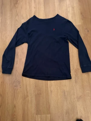 Marinblå långärmad topp Polo Ralph Lauren - Klassisk marinblå långärmad topp från Polo Ralph Lauren med rund halsringning och diskret röd logga broderad på bröstet. Tillverkad i mjuk bomull som känns skön mot huden. Perfekt till jeans eller kjol för en clean och tidlös look.