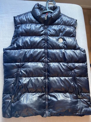 Mörkblå dunväst från Moncler - Snygg mörkblå dunväst från Moncler med hög krage och två dragkedjeförsedda fickor. Västen har ett quiltat mönster och klassisk Moncler-logga på bröstet. Perfekt för lager-på-lager och håller dig varm under kyliga dagar. Inte äkta 