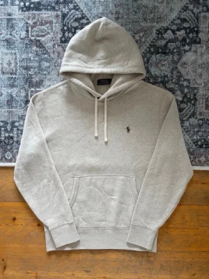 Beige hoodie från Polo Ralph Lauren - Snygg beige hoodie från Polo Ralph Lauren med klassisk huva och dragsnören. Har en broderad logga på bröstet och en stor magficka framtill. Tillverkad i mjukt material och har ribbade muddar vid ärmar och nederkant. Nytväddad när man får hem den, även den enda gången den blivit tvättad 