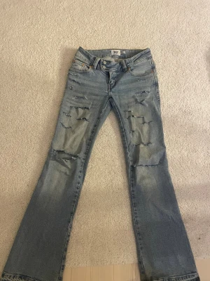 Ljusblå slitna bootcut jeans - Säljer ett par ljusblå bootcut jeans med slitningar och hål på framsidan. Jeansen har låg midja och broderade detaljer på bakfickorna. Tillverkade i denim med en klassisk tvättad look.