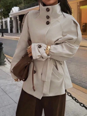 Beige trenchcoat med hög krage - Stilren beige trenchcoat med hög krage och stora knappar framtill. Jackan har ett knytskärp i midjan, breda ärmar med knappdetaljer och stora fickor. Så snygg nu till hösten😍Helt oanvänd, satt för stort på mig