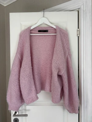 Ljusrosa stickad kofta Vero Moda - Mysig ljusrosa stickad kofta från Vero Moda med oversized passform och långa ärmar. Koftan är öppen framtill och har ribbade muddar vid ärmslut och nederkant. Perfekt för lager på lager och ger en soft vibe till din outfit.