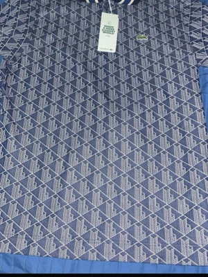 Blå mönstrad piké från Lacoste - Snygg blå pikétröja från Lacoste med vitt geometriskt mönster och klassisk krage med vita ränder. Tröjan har knappslå framtill och den ikoniska krokodilloggan på bröstet. Perfekt för dig som gillar sportig och stilren look.