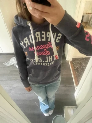 Mörkgrå Superdry hoodie med tryck - Säljer en mörkgrå hoodie från Superdry med stora vita och rosa tryck på bröstet och ärmen. Hoodien är väldigt trendig och eftertraktad! 