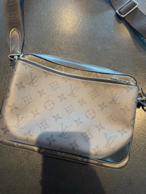   Svart axelväska från Louis Vuitton - Snygg svart axelväska från Louis Vuitton med klassiskt monogrammönster. Väskan har justerbar axelrem i textil och detaljer i silverfärgad metall. Tillverkad i läder med dragkedja upptill och stilren design.