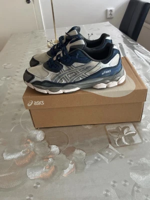 Asics sneakers i blått och grått - Snygga Asics sneakers i blått, grått och vitt med klassisk mesh och syntetmaterial. Skorna har lågt skaft, tydliga Asics-loggor på sidan och en chunky sula som ger en sportig vibe. Perfekta för dig som gillar streetwear och bekväma skor.