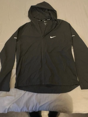 Grå Nike miller - Snygg svart vindjacka från Nike med hel dragkedja och huva. Jackan har reflekterande detaljer på ärmarna och bröstet samt lång ärm. Tillverkad i lätt och tunt material, perfekt för blåsiga dagar. Klassisk Nike-logga på bröstet.