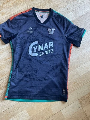Svart NOCTA fotbollströja - Snygg svart fotbollströja från NOCTA med trycket 'CYNAR SPRITZ' på bröstet. Tröjan har färgglada detaljer i rött och grönt längs axlar och sidor, samt ett diskret mönster över hela tyget. Perfekt för träning eller match.