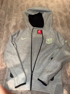 Grå Nike FC Barcelona hoodie - Snygg grå hoodie från Nike med FC Barcelona-logga på bröstet och limegrön swoosh. Tröjan har dragkedja, huva och svarta detaljer vid fickorna. Perfekt för dig som gillar sportig stil och vill visa vilket lag du hejar på.
