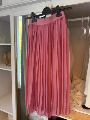Plisserad rosa långkjol - Säljer en snygg plisserad långkjol i rosa med bred linning och ett litet V-snitt framtill. Kjolen har ett luftigt fall och är perfekt för dig som vill ha en feminin och trendig look. Materialet känns lätt och flowy.
