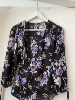 Svart blommig omlottblus - Snygg svart omlottblus med lila, vita och blå blommor. Blusen har långa ärmar med resår vid ärmsluten och knytband i midjan för en feminin siluett. Perfekt för dig som gillar romantiska mönster och mjuka material.