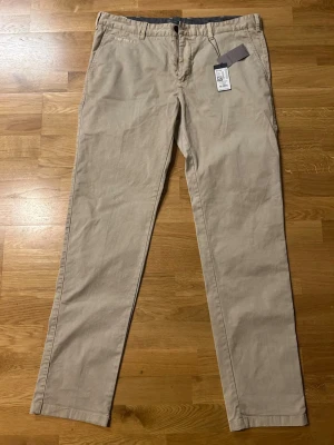 Beige chinos Marc O'Polo - Snygga beige chinos från Marc O'Polo i rak modell. Byxorna har klassiska fickor fram och bak, bälteshällor och stängs med knapp och dragkedja. Tillverkade i ett mjukt bomullsmaterial som ger en clean och stilren look.