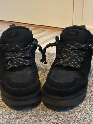 Svarta sneakers från UGG - Snygga svarta sneakers från UGG med chunky sula och breda, mönstrade skosnören. Ovandelen är i mocka och mesh för en cool och sportig look. Perfekta för dig som gillar streetwear och vill ha något bekvämt och trendigt.