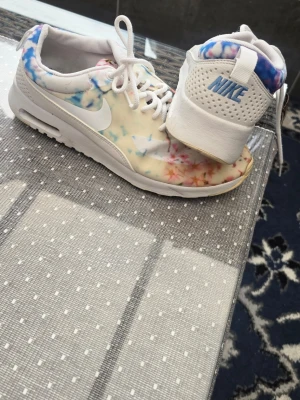 Nike sneakers med blommigt mönster - Nike sneakers med vit bas och färgglatt blommigt mönster i pastellfärger. Skorna har vita snören, vit sula och en klassisk Nike-logga på sidan. Lätta och snygga sneakers med låg profil, perfekta för en trendig streetstyle.