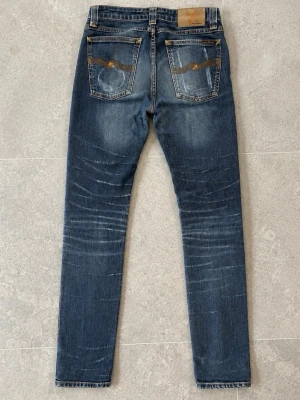 Nudie Jeans 28/34 - Nudie jeans med tydliga slitningar och whiskers på både fram- och baksida. Jeansen har klassiska fem fickor och kontrastsömmar. Materialet är denim i bomull med stretch för extra komfort. Snygga detaljer på bakfickorna.