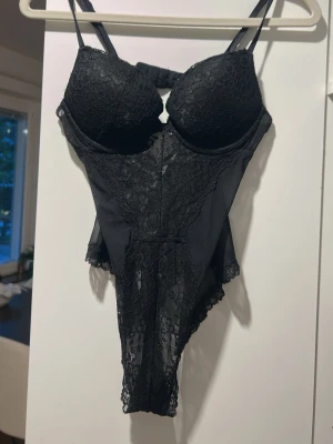 Svart spetsbody med tunna axelband från H&M - Snygg svart body i spets med tunna justerbara axelband och vadderade kupor. Framsidan har dekorativa spetsdetaljer och baksidan är i transparent mesh med en liten öppning. Perfekt för dig som vill ha något både elegant och bekvämt.