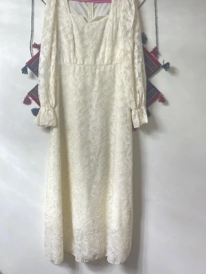 Lång vit spetsklänning med puffärm - Elegant lång klänning i vit spets med blommigt mönster. Klänningen har långa puffärmar med volang vid ärmslut och en markerad midja. V-ringad halsringning och dragkedja bak. Perfekt för dig som gillar vintage vibes och romantisk stil.