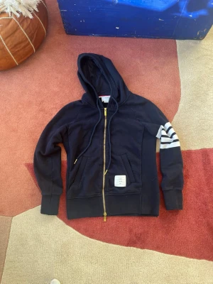 Mörkblå hoodie med vita ränder ”Thom browne ” Size 0 - Snygg mörkblå hoodie med dragkedja och huva. Har fyra vita ränder på ena ärmen och en patch med text på magen. Guldiga detaljer på dragkedjan och två fickor framtill. Perfekt för en avslappnad och trendig stil.