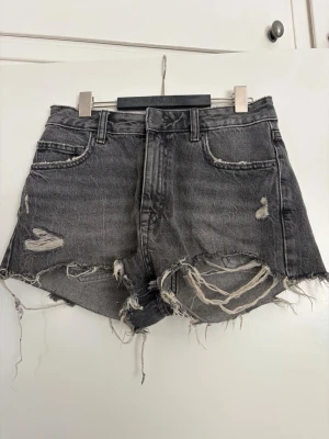 Gråa slitna denimshorts - Säljer ett par gråa midwaist jeansshorts med råa kanter och slitningar framtill. Shortsen har fem fickor, bälteshällor och dragkedja med knapp. Perfekta för en avslappnad och edgy stil.