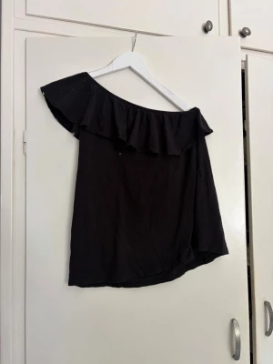 Svart offshoulder topp med volang - Svart offshoulder topp från L.O.G.G. med bred volang längs halsringningen. Toppen är ärmlös och har en snygg, asymmetrisk design som ger en trendig vibe. Perfekt att matcha med jeans eller kjol för en cool look.