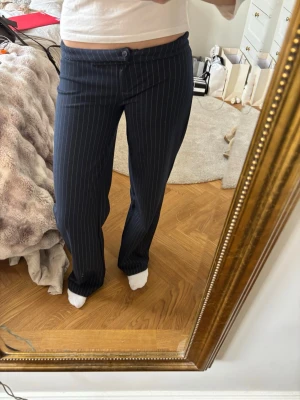 Mörkblå randiga kostymbyxor - Säljer ett par mörkblå  kostymbyxor med vita tunna streck. Byxorna har en mer baggy passform, lågmidjade och stängs med knapp och dragkedja. 