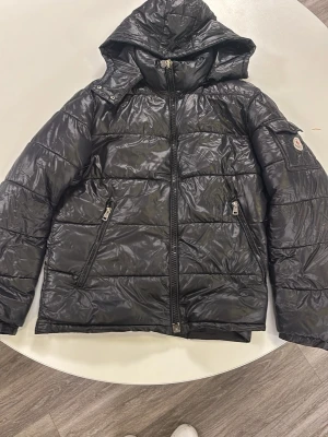 Moncler Maja  - Snygg svart dunjacka från Moncler med huva och två dragkedjeförsedda fickor framtill. Jackan har quiltad design och Moncler-logga på ärmen. Perfekt för kalla vinterdagar och riktigt stilren look.