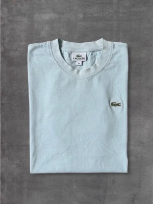 Ljusblå t-shirt från Lacoste - Snygg ljusblå t-shirt från Lacoste med klassisk rund hals och det ikoniska krokodilmärket broderat på bröstet. Tillverkad i mjuk bomull och har en normal passform. Perfekt för en clean och stilren look.