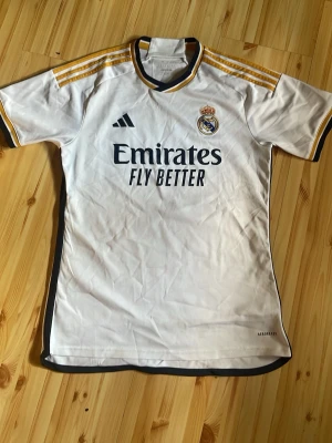 Real Madrid matchtröja Adidas M - Säljer en vit Real Madrid matchtröja från Adidas med guldfärgade detaljer på axlarna och klubbmärke på bröstet. Tröjan har svart Adidas-logga och texten 'Emirates Fly Better' framtill. Tillverkad i lätt och ventilerande material, perfekt för fotboll.