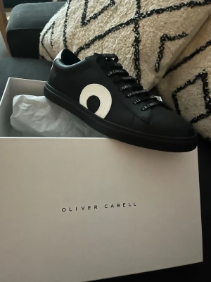 Oliver Cabell svarta sneakers low - Snygga svarta sneakers från Oliver Cabell med vit cirkeldetalj på sidan och svarta skosnören med logga. Modellen är low och färgen heter Daybreak. Tillverkade i läder för en clean och stilren look. Helt nya 