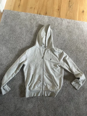 Ljusgrå hoodie Ralph lauren  - Snygg ljusgrå hoodie med dragkedja framtill och två stora fickor. Den är i storlek S men passar m. Klassisk huva med snörning och diskret broderad logga på bröstet. Tillverkad i mjukt sweatshirtmaterial.